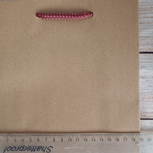 May include: A brown paper gift bag with red rope handles. The bag is sitting on a wooden surface with a ruler underneath.