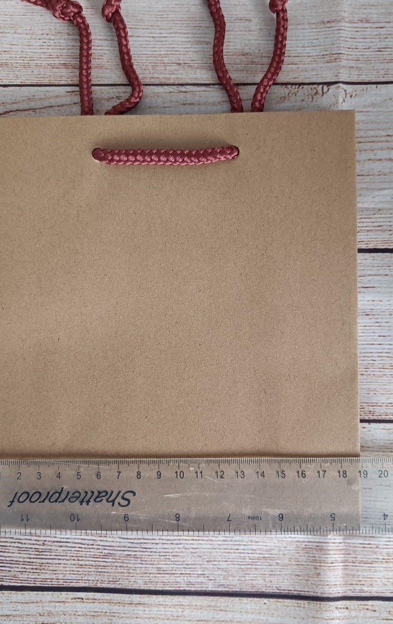 May include: A brown paper gift bag with red rope handles. The bag is sitting on a wooden surface with a ruler underneath.