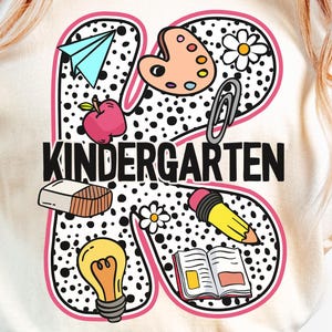 May include: A cream-colored t-shirt featuring a large letter K with a black and white polka dot pattern. The letter is decorated with colorful school-themed icons including a paper airplane, paint palette, apple, paperclip, eraser, lightbulb, pencil, and open book, with the word KINDERGARTEN printed in black.
