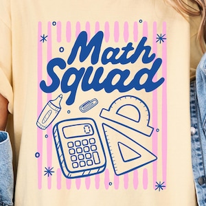 May include: A pale yellow t-shirt featuring a graphic print with the text "Math Squad" in blue. The design includes illustrations of a calculator, a protractor, a triangle ruler, a paperclip, and a marker, all set against a background of vertical pink stripes.