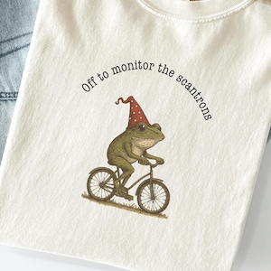 May include: Cream-colored t-shirt with a frog riding a bicycle, wearing a party hat. The text "Off to monitor the scantrons" is printed above the frog. The shirt is paired with blue jeans and a tan hat.