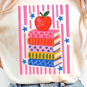 May include: An ivory t-shirt featuring a graphic print of a stack of five colorful books topped with a red apple. The design includes vertical pink stripes and blue stars surrounding the books. The shirt has short sleeves and a crew neckline.