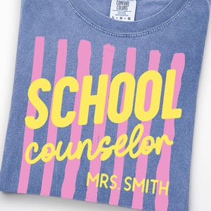 May include: A blue t-shirt featuring a graphic of vertical pink stripes with the yellow text SCHOOL counselor and MRS. SMITH printed over the stripes. The shirt has a crew neckline and a Comfort Colors brand label visible at the inner collar.