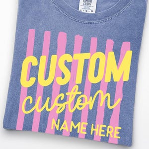 May include: A blue short-sleeved t-shirt with a Comfort Colors label at the neck. The front features vertical pink brushstroke stripes with the yellow text CUSTOM, custom, and NAME HERE printed over them. A circular logo reads COMFORT COLORS BLUE JEAN.