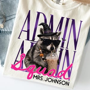 May include: An ivory t-shirt featuring a graphic of a raccoon wearing a black witch hat, sunglasses, and a beaded necklace while holding an iced coffee. The text reads ADMIN in purple, Squad in pink, and MRS. JOHNSON in black below the raccoon.
