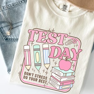 May include: Cream-colored t-shirt with a retro "Test Day" graphic in pink and blue. The design includes pencils, an apple, and books. The shirt is paired with light-wash jeans and a tan hat.