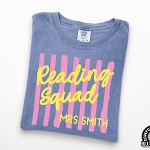May include: A blue jean colored t-shirt with a crew neckline and a Comfort Colors label. The front features vertical pink brushstroke stripes with the yellow text Reading Squad and the name MRS. SMITH printed in a bold yellow sans-serif font.