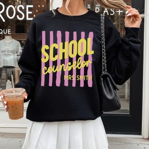 May include: A black long-sleeve crewneck sweatshirt featuring a graphic design with vertical pink brushstroke stripes. The text reads SCHOOL counselor in yellow, with MRS. SMITH printed below in yellow. The sweatshirt has a ribbed neckline.