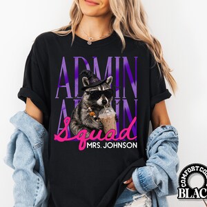 May include: A black t-shirt featuring a graphic of a raccoon wearing a witch hat, sunglasses, and a pumpkin bead necklace while holding an iced coffee. The text reads ADMIN SQUAD and MRS. JOHNSON in purple and pink lettering on the front.