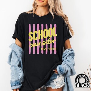 May include: A black short-sleeve t-shirt featuring a graphic design with vertical pink brushstroke stripes. The text SCHOOL COUNSELOR is printed in yellow, with MRS. SMITH printed below in yellow. The shirt has a crew neckline and short sleeves.