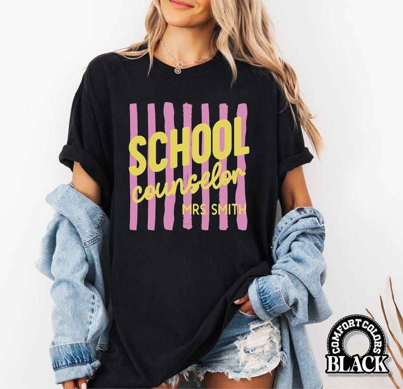 May include: A black short-sleeve t-shirt featuring a graphic design with vertical pink brushstroke stripes. The text SCHOOL COUNSELOR is printed in yellow, with MRS. SMITH printed below in yellow. The shirt has a crew neckline and short sleeves.