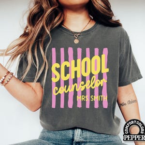 May include: A charcoal gray short-sleeve t-shirt featuring a graphic design of vertical pink stripes. The text SCHOOL counselor MRS. SMITH is printed in yellow over the stripes. The shirt has a crew neckline and a relaxed, vintage-washed appearance.