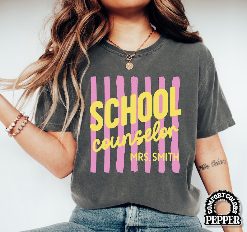 May include: A charcoal gray short-sleeve t-shirt featuring a graphic design of vertical pink stripes. The text SCHOOL counselor MRS. SMITH is printed in yellow over the stripes. The shirt has a crew neckline and a relaxed, vintage-washed appearance.