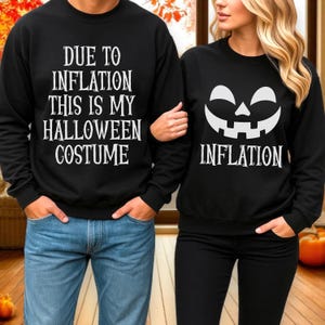 May include: Two black crewneck sweatshirts. The left sweatshirt features white text reading DUE TO INFLATION THIS IS MY HALLOWEEN COSTUME. The right sweatshirt displays a white jack-o'-lantern face graphic above the word INFLATION in white text.