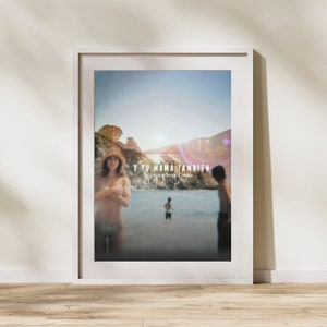 May include: Framed movie poster for "Y Tu Mamá También" featuring a beach scene with three people. The poster has a white frame and is displayed against a white wall. The movie title is at the top.