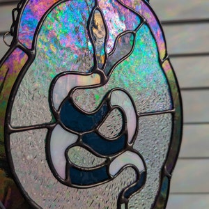 May include: A stained glass window hanging with a black metal frame and iridescent glass. The design features a white snake with a blue outline, curled in a spiral shape.