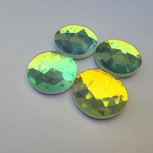 May include: Four iridescent, faceted, round gemstones. The gems have a mix of green and yellow hues, with a white backing. The gems are arranged in a circular pattern against a light gray background. These could be used for jewelry making or craft projects.