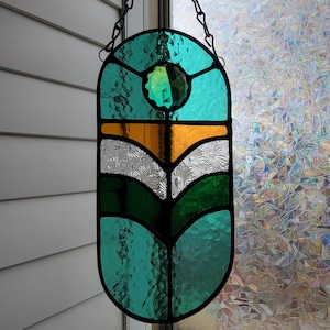 May include: A stained glass window hanging from a chain. The window is oval shaped and features a green, white, and gold geometric design with a green gem in the center.