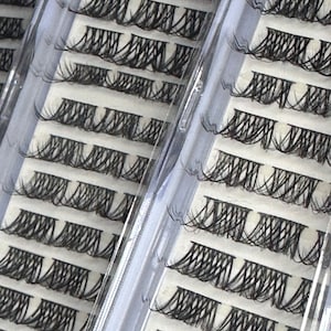Pode incluir: Close-up de vários conjuntos de cílios postiços pretos, cuidadosamente dispostos em bandejas de plástico transparente. Cada bandeja contém várias fileiras de tufos de cílios individuais, mostrando uma variedade de comprimentos e estilos para maquiagem dos olhos.