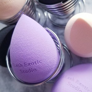 May include: A collection of makeup sponges in various colors, including lavender, black, and peach. Each sponge is held in a silver wire holder. The text "Lash Exotic Studio" is printed on some of the sponges.