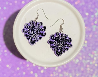 May include: A pair of purple and black earrings with a decorative design, displayed on a white plate against a sparkly purple background. The earrings have a floral-like shape with a detailed black pattern.