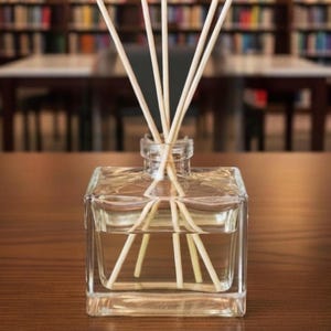May include: A clear, square glass diffuser bottle with several light-coloured reeds. The bottle sits on a wooden surface, with a blurred background of bookshelves and tables. The reeds are arranged to fan out from the bottle's opening.