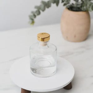 Pode incluir: Uma garrafa de vidro transparente com tampa dourada, cheia de líquido transparente. A garrafa está sobre um suporte redondo de madeira branco. Um vaso de madeira e vegetação ao fundo.