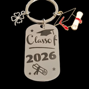 May include: Silver keychain with a graduation theme. The keychain features a dog tag engraved with "Class of 2026", a graduation cap charm, a diploma charm, and a four-leaf clover charm. The background is black.