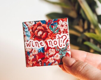 May include: Square magnet with a floral pattern in red, white, and blue. The magnet has the words "wine not?" in white script. The magnet is held by a hand.