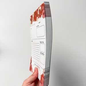 May include: A small, white, floral-patterned notebook with a spiral binding. The notebook has sections for "date", "priorities", "things to buy", and "until next time".