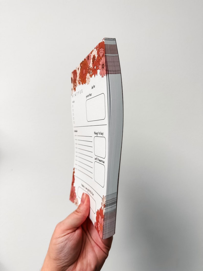 May include: A small, white, floral-patterned notebook with a spiral binding. The notebook has sections for "date", "priorities", "things to buy", and "until next time".