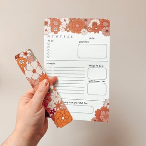 May include: A white planner with a floral border in shades of orange and pink. The planner has sections for daily tasks, priorities, schedule, things to buy, and a gratitude section. The planner is held by a hand.