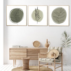 K&ouml;nnte beinhalten: Drei gerahmte Drucke mit minimalistischen Linienzeichnungen verschiedener Pflanzen auf hellgr&uuml;nem Hintergrund. Die Drucke h&auml;ngen an einer wei&szlig;en Wand &uuml;ber einer Kommode aus Holz mit einem geflochtenen Korb und einem Stuhl mit einem Strohhut.