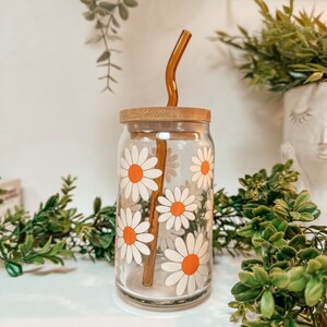 Pu&ograve; includere: Una bottiglia d'acqua riutilizzabile in vetro trasparente con un coperchio in bamb&ugrave; e una cannuccia in vetro marrone. La bottiglia ha un motivo di margherita bianca con centri arancioni.