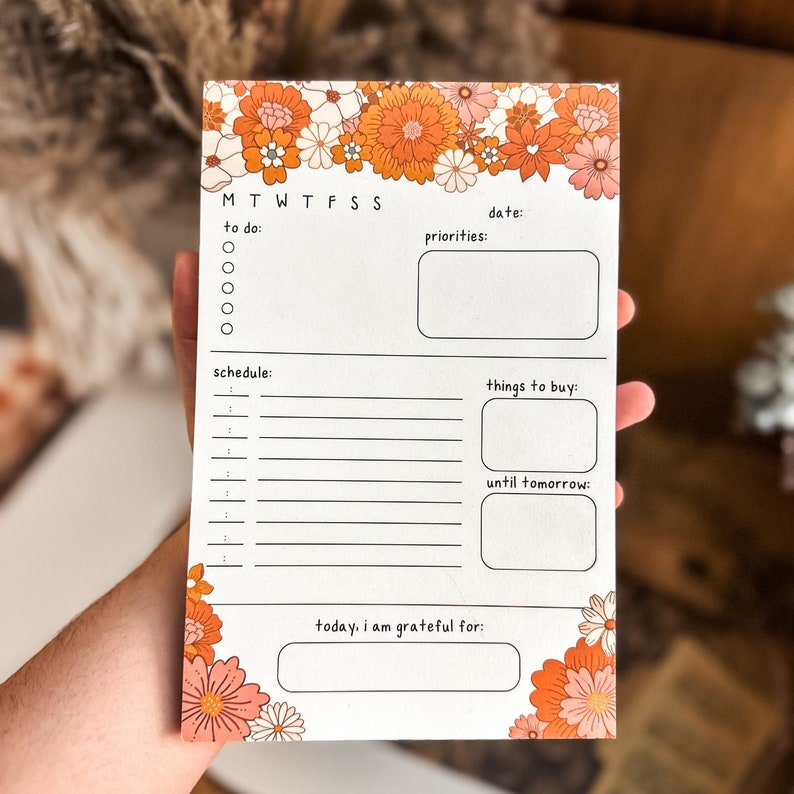 May include: A white planner with a floral border featuring orange, pink, and white flowers. The planner has sections for to do, priorities, schedule, things to buy, and until tomorrow. It also has a section for "today, I am grateful for".