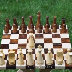 May include: A wooden chess set with brown and white pieces on a wooden chessboard with a brown and white checkered pattern. The chessboard is on a wooden table with a green grassy background. The text "Chess'n'Boards" is visible on the board.