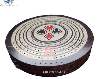 May include: A wooden cribbage board with a circular design. The board has a cream-colored surface with black markings and a center circle featuring playing card suits. The board has a brown wooden frame and the text "GAMES WON" is printed on the board.