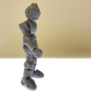 Puede incluir: Figura de robot articulado gris con un dise&ntilde;o detallado. El robot tiene una cabeza redondeada, brazos y piernas articulados y una expresi&oacute;n ligeramente triste. La figura est&aacute; de pie sobre una superficie blanca contra un fondo claro.