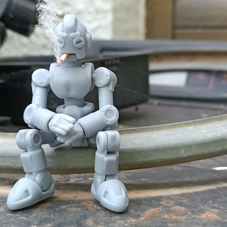 Puede incluir: Una figura de robot gris articulada est&aacute; sentada, con un cigarrillo encendido en la boca y humo ascendente. El robot tiene un dise&ntilde;o mec&aacute;nico con articulaciones visibles y detalles circulares. La figura descansa sobre una superficie oscura.