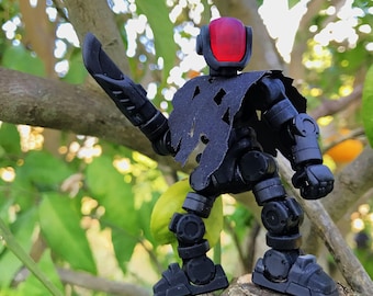Puede incluir: Una figura de acción negra articulada con una visera roja y una capa rasgada, sosteniendo una espada negra. La figura posa sobre una rama de árbol contra un fondo borroso de hojas verdes y fruta.