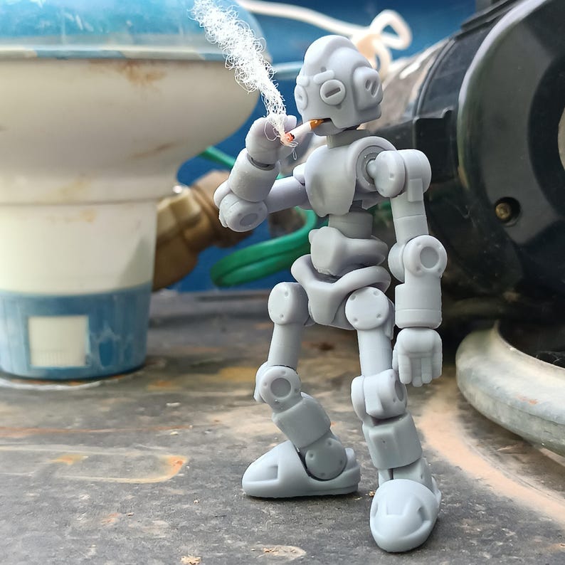 Puede incluir: Una figura de robot gris articulada sosteniendo un cigarrillo encendido, con humo ascendente. El robot tiene un dise&ntilde;o mec&aacute;nico con articulaciones visibles y una cabeza redondeada. El fondo incluye un objeto azul y blanco y una estructura cil&iacute;ndrica negra.