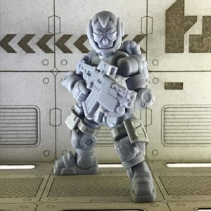 Può includere: Action figure in plastica grigia di un soldato futuristico, che tiene un fucile. La figura è molto dettagliata, con armature e un elmetto. Lo sfondo è una superficie metallica con segni industriali.