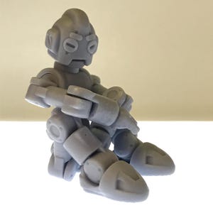 Puede incluir: Una figura de robot articulada gris con cabeza redondeada, ojos ovalados y extremidades articuladas. El robot est&aacute; en posici&oacute;n sentada, con los brazos cruzados y las piernas dobladas. La figura est&aacute; hecha de un material liso y s&oacute;lido.