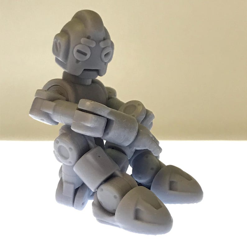 Puede incluir: Una figura de robot articulada gris con cabeza redondeada, ojos ovalados y extremidades articuladas. El robot est&aacute; en posici&oacute;n sentada, con los brazos cruzados y las piernas dobladas. La figura est&aacute; hecha de un material liso y s&oacute;lido.