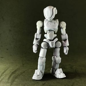May include: A light gray articulated robot action figure with a rounded helmet, shoulder pads, and jointed limbs. The robot has a chest plate with the letters "OKI" and is standing on a green surface. The figure is designed with multiple points of articulation.