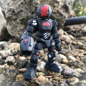 Op de afbeelding: Een zwart gearticuleerd robotactiefiguur met rode en witte accenten. De robot heeft een rood frontpaneel, witte markeringen en het woord "OU" op de borst. Het staat op een rotsachtig oppervlak.