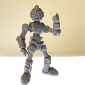 Puede incluir: Una figura de robot articulada gris con una cabeza redondeada, ojos circulares y una mano que se&ntilde;ala hacia arriba. El robot tiene m&uacute;ltiples articulaciones, lo que permite varias poses. La figura est&aacute; sobre una superficie blanca.
