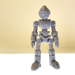 Puede incluir: Una figura de robot articulada gris con cabeza redondeada, ojos circulares y extremidades articuladas. La figura est&aacute; de pie, con los brazos a los lados. El dise&ntilde;o del robot es simple y geom&eacute;trico, con un acabado liso y mate.