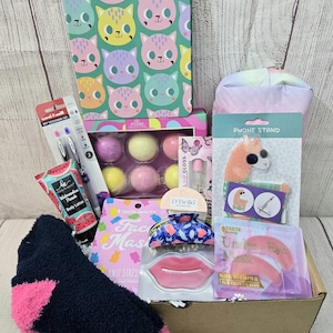 May include: A gift box filled with self-care items. Includes a cat-themed notebook, bath bombs, lip gloss, a phone stand, face mask, hair clip, lip mask, under-eye masks, a pen, body lotion, and a navy blue and pink sock.