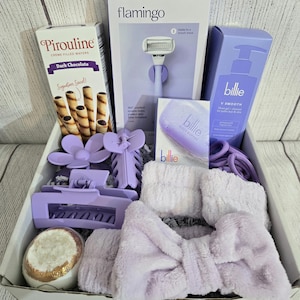 May include: A gift box filled with self-care items in shades of purple and white. Includes a razor, shave gel, bath bomb, hair accessories, a headband, and dark chocolate creme-filled wafers. The box is presented on a light wood-grain background.